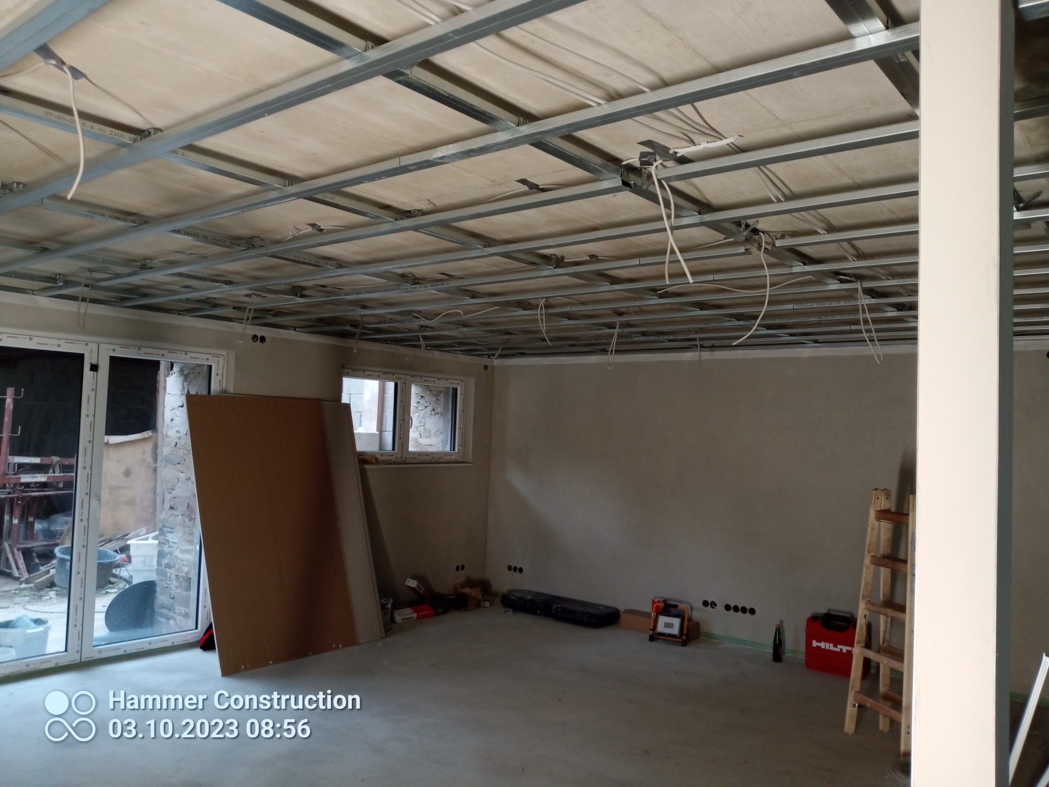 Wnętrze w trakcie montażu metalowej konstrukcji sufitu podwieszanego z widocznymi przewodami, niezabezpieczonymi ścianami i elementami wyposażenia budowlanego w tle.