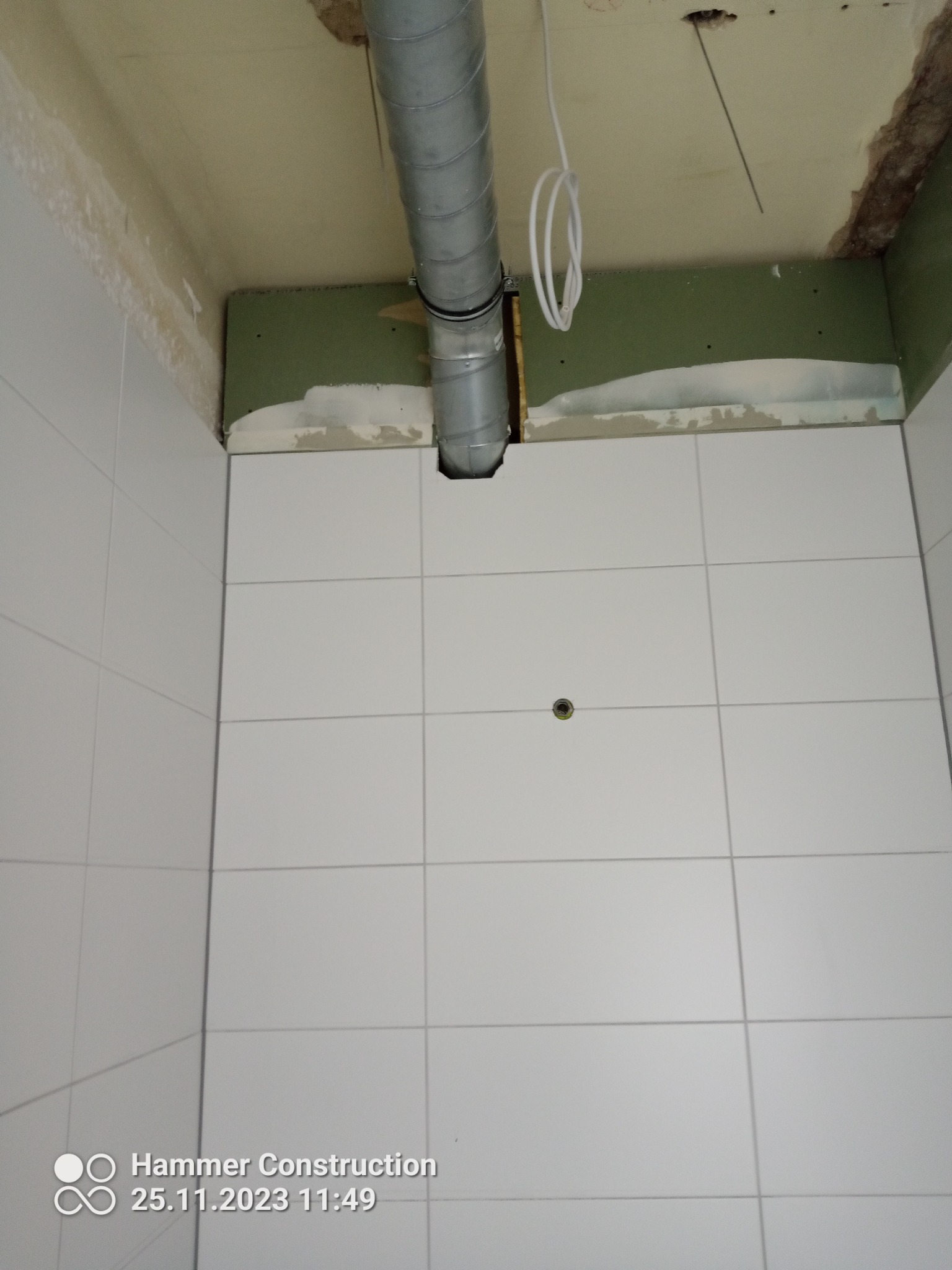 Fragment łazienki w trakcie prac: białe płytki ścienne, widoczny fragment sufitu z wystającą rurą wentylacyjną i fragmentem płyty gipsowej.