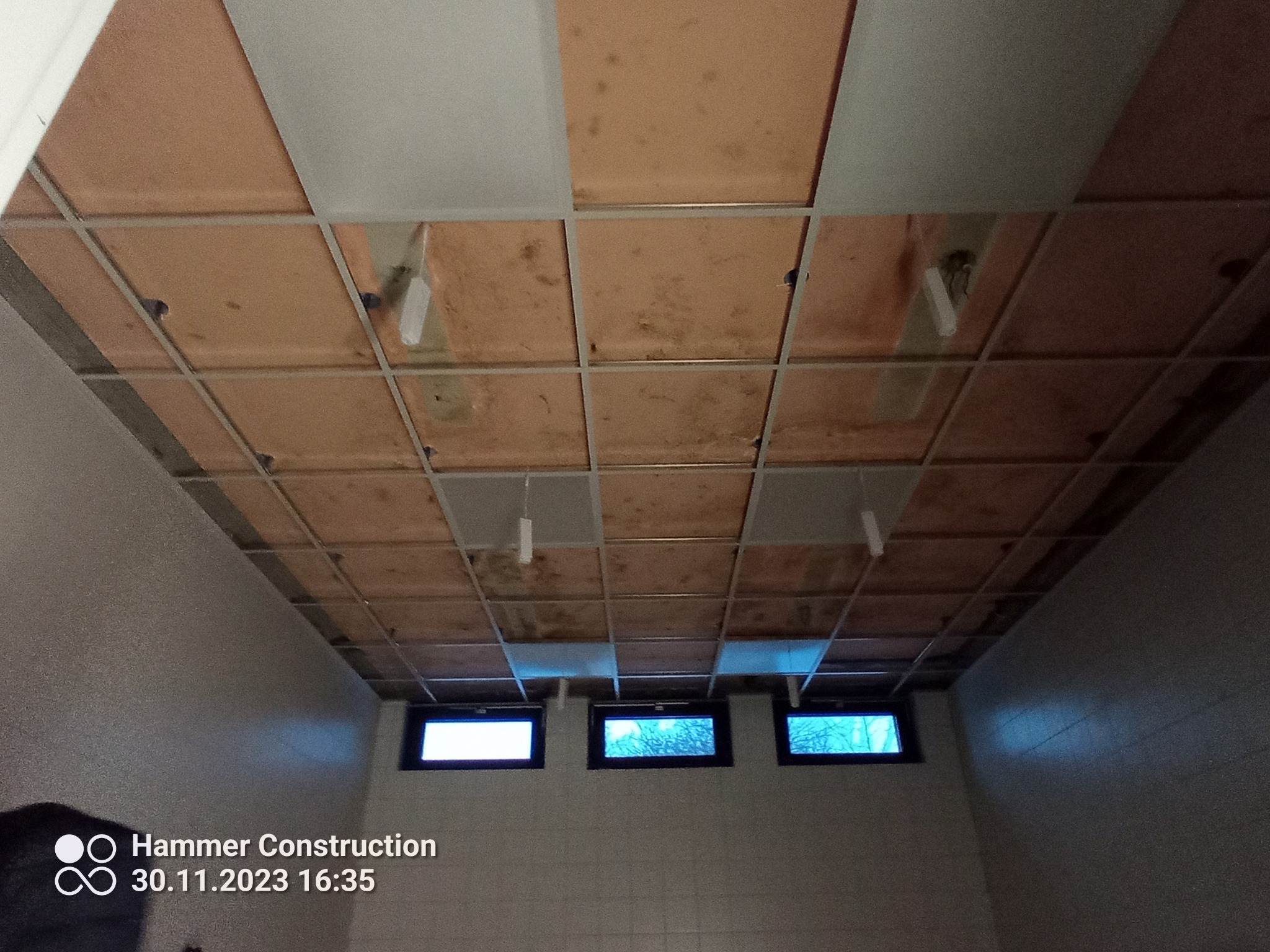 Widok na sufit podwieszany z widoczną konstrukcją metalową, częściowo zamontowanymi panelami i instalacją oświetleniową w pomieszczeniu z trzema małymi oknami.