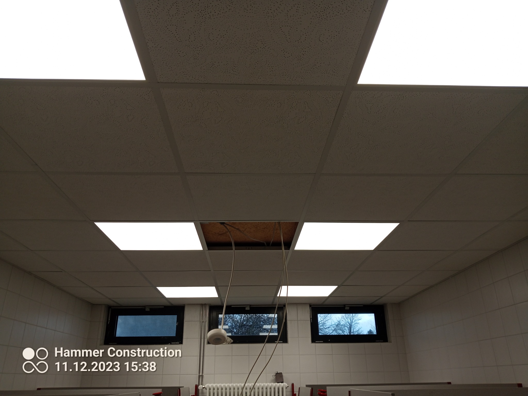 Sufit podwieszany z usuniętą płytą, widoczne okablowanie i fragment instalacji nad sufitem, w tle okna.