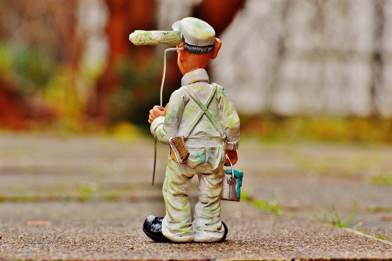 Figurka malarza z wałkiem na głowie, pędzlem w kieszeni i wiaderkiem farby, stojąca na chodniku.
