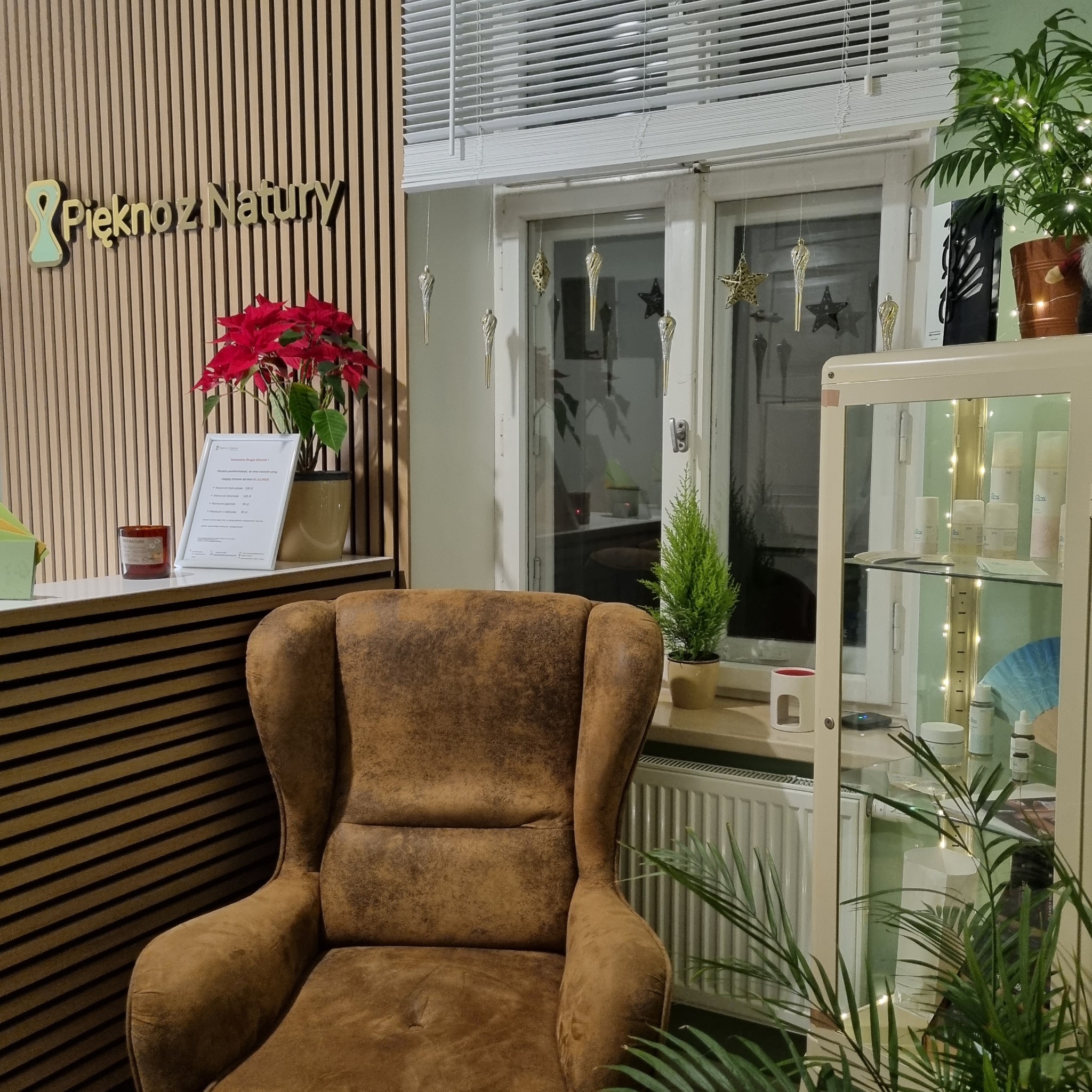 Wnętrze gabinetu z fotelem, kwiatami i witryną z kosmetykami, w tle okno z dekoracjami i logo firmy 'Pięknoz Natury'.