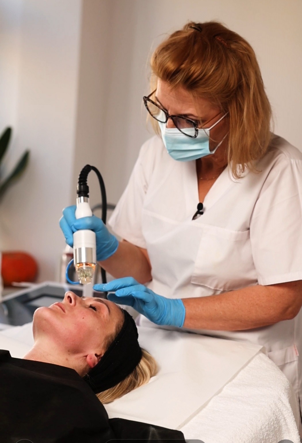 Kobieta w masce i okularach wykonuje zabieg na twarzy klientki za pomocą specjalistycznego urządzenia z igłami.