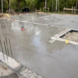 Świeżo wylana płyta fundamentowa z wystającymi prętami zbrojeniowymi, widoczne elementy instalacji oraz przygotowane miejsce na wkop z piaskiem i izolacją, w tle las.