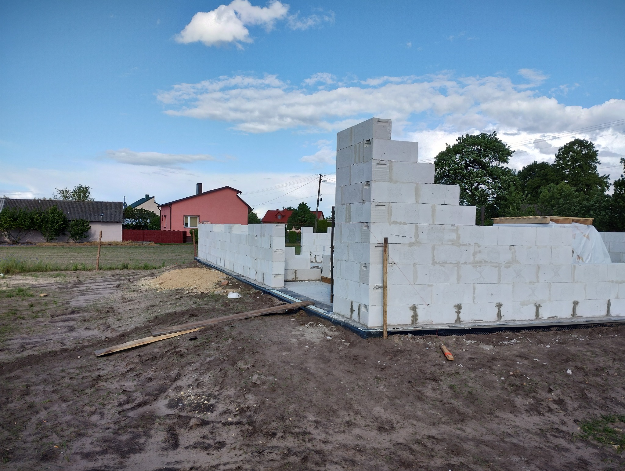 Wznoszone ściany domu z białych bloczków betonowych na fundamencie z widoczną izolacją przeciwwilgociową, w tle zabudowa jednorodzinna i błękitne niebo z chmurami.
