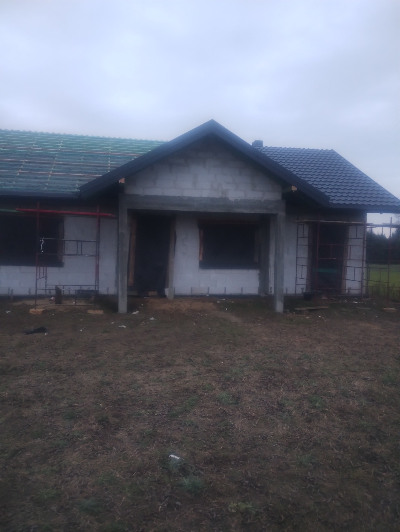 Niedokończony budynek z szarych bloczków, z częściowo położonym zielonym dachem z jednej strony i czarną dachówką z drugiej, otoczony rusztowaniami i brązową ziemią.