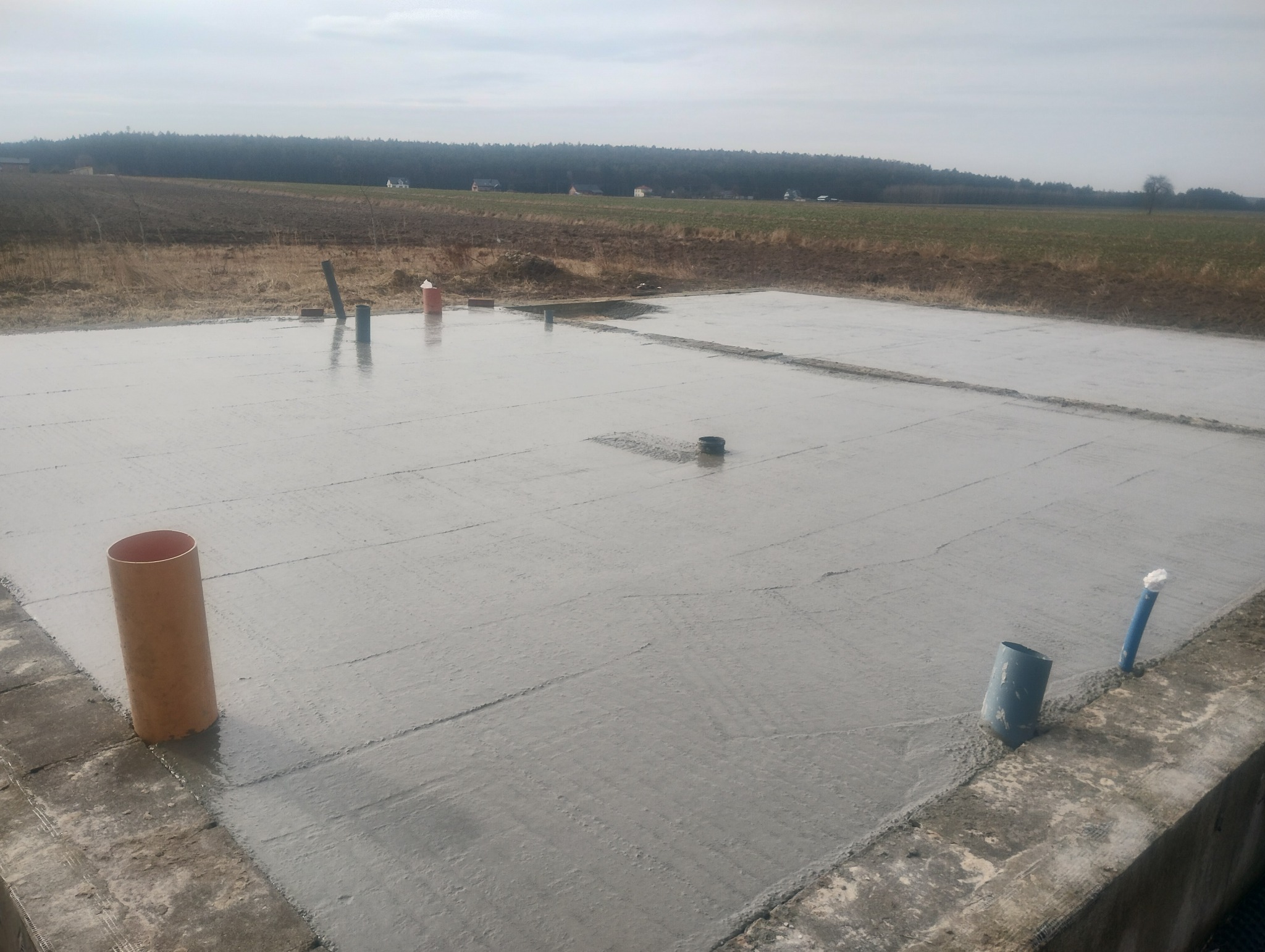 Świeżo wylana płyta fundamentowa na terenie budowy w Bałtowie z widocznymi pionowymi rurami instalacyjnymi i odległym polem uprawnym w tle.