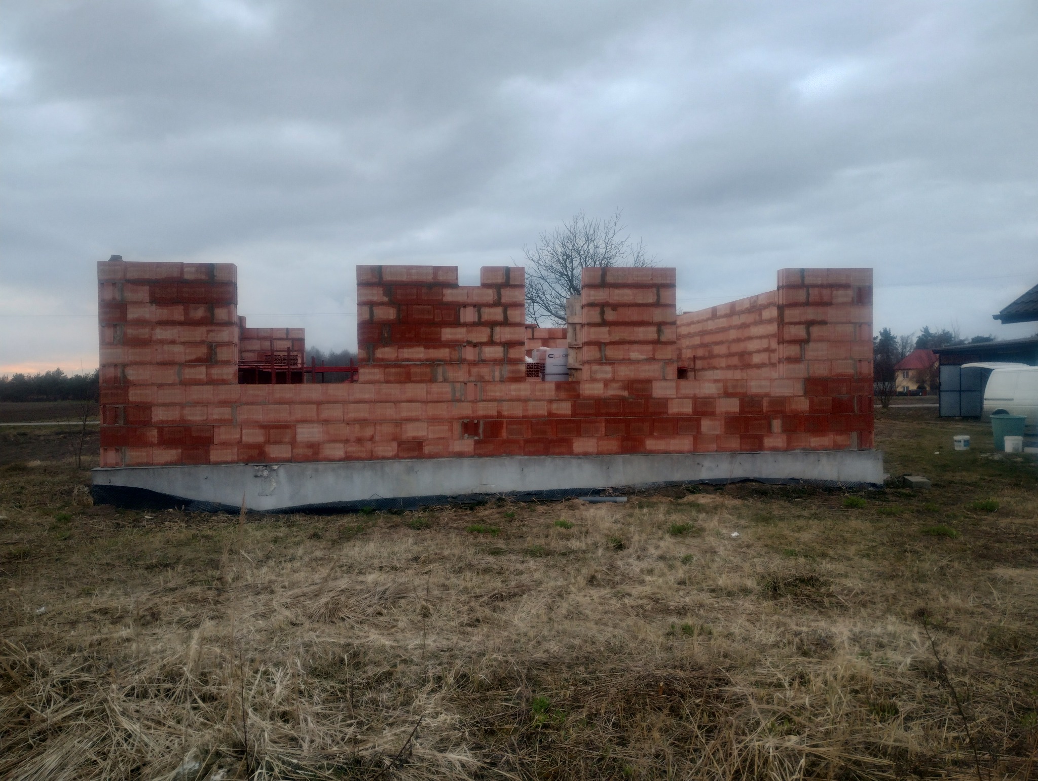 Wznoszone ściany domu z czerwonej cegły na wylanym fundamencie, widok na plac budowy w pochmurny dzień.