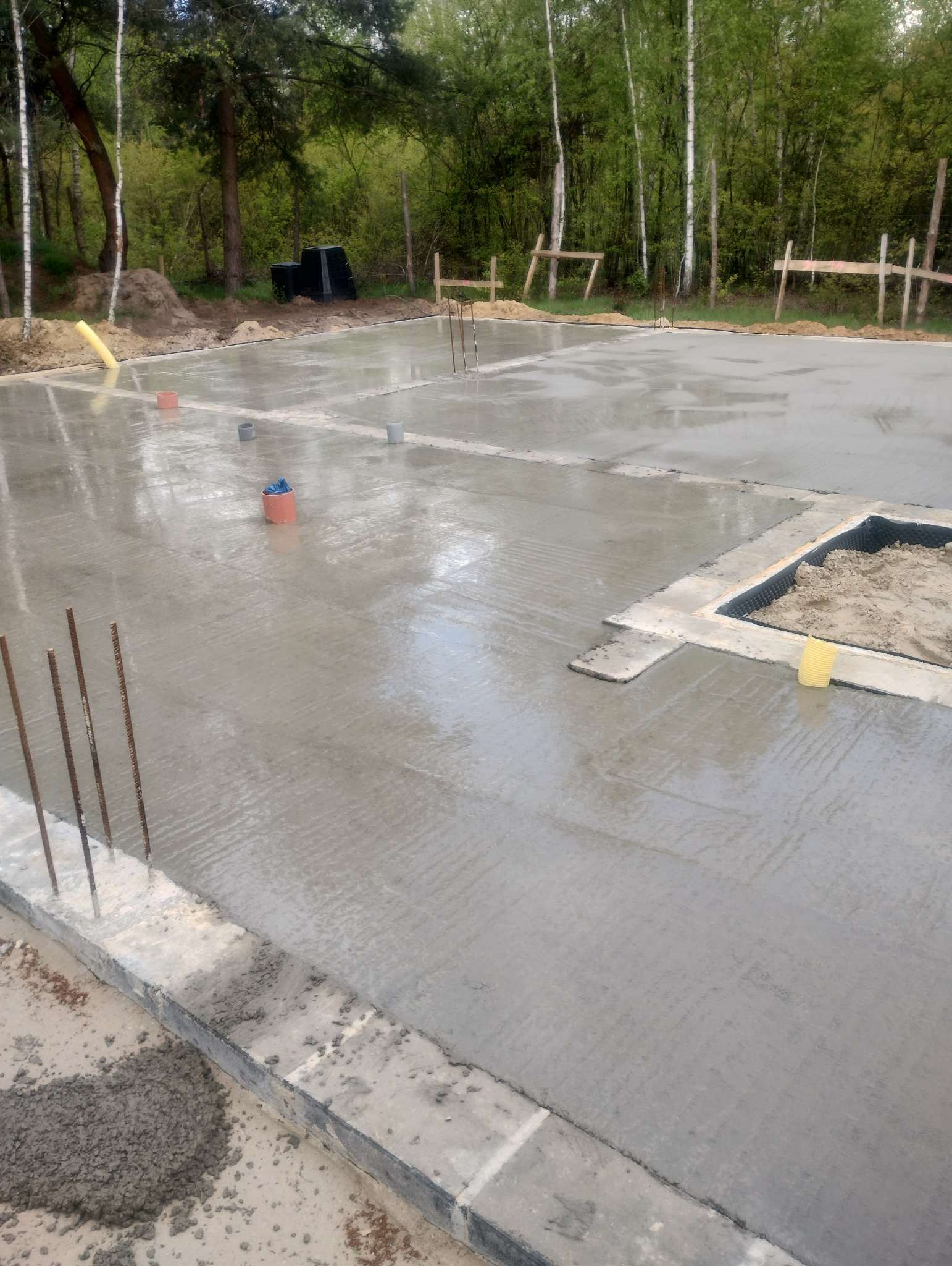 Świeżo wylana płyta fundamentowa z wystającymi prętami zbrojeniowymi, widoczne elementy instalacji oraz przygotowane miejsce na wkop z piaskiem i izolacją, w tle las.