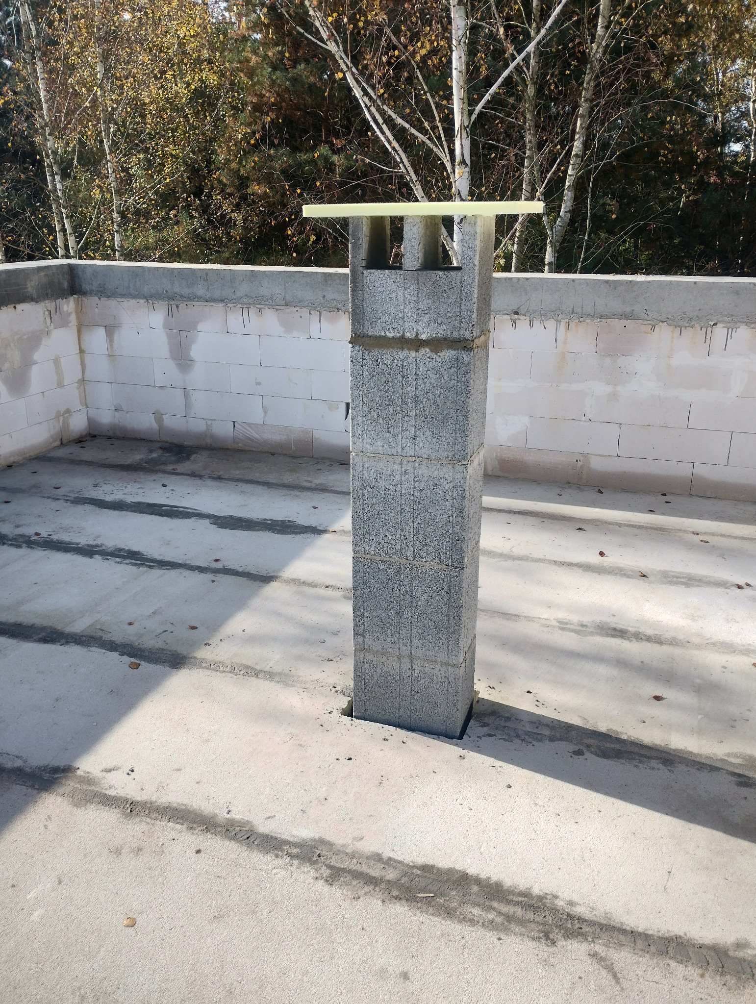 Wzniesiony komin z bloczków betonowych na stropie budynku w trakcie budowy, widoczne ślady po deskowaniu i białe ściany z bloczków silikatowych w tle.