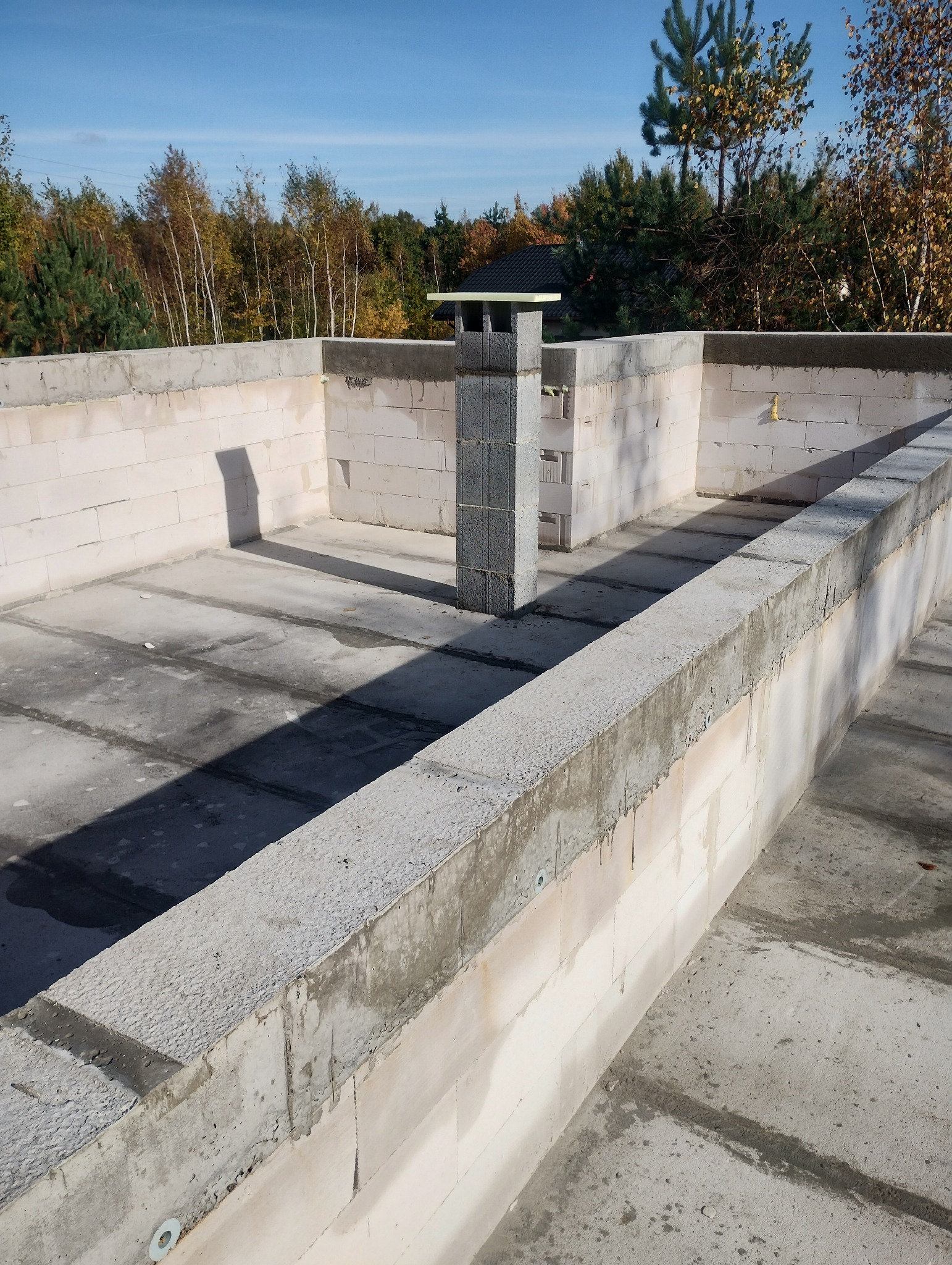 Wnętrze domu w trakcie budowy: ściany z białych bloczków, wylany betonowy fundament, komin z szarych bloczków na tle jesiennego krajobrazu.