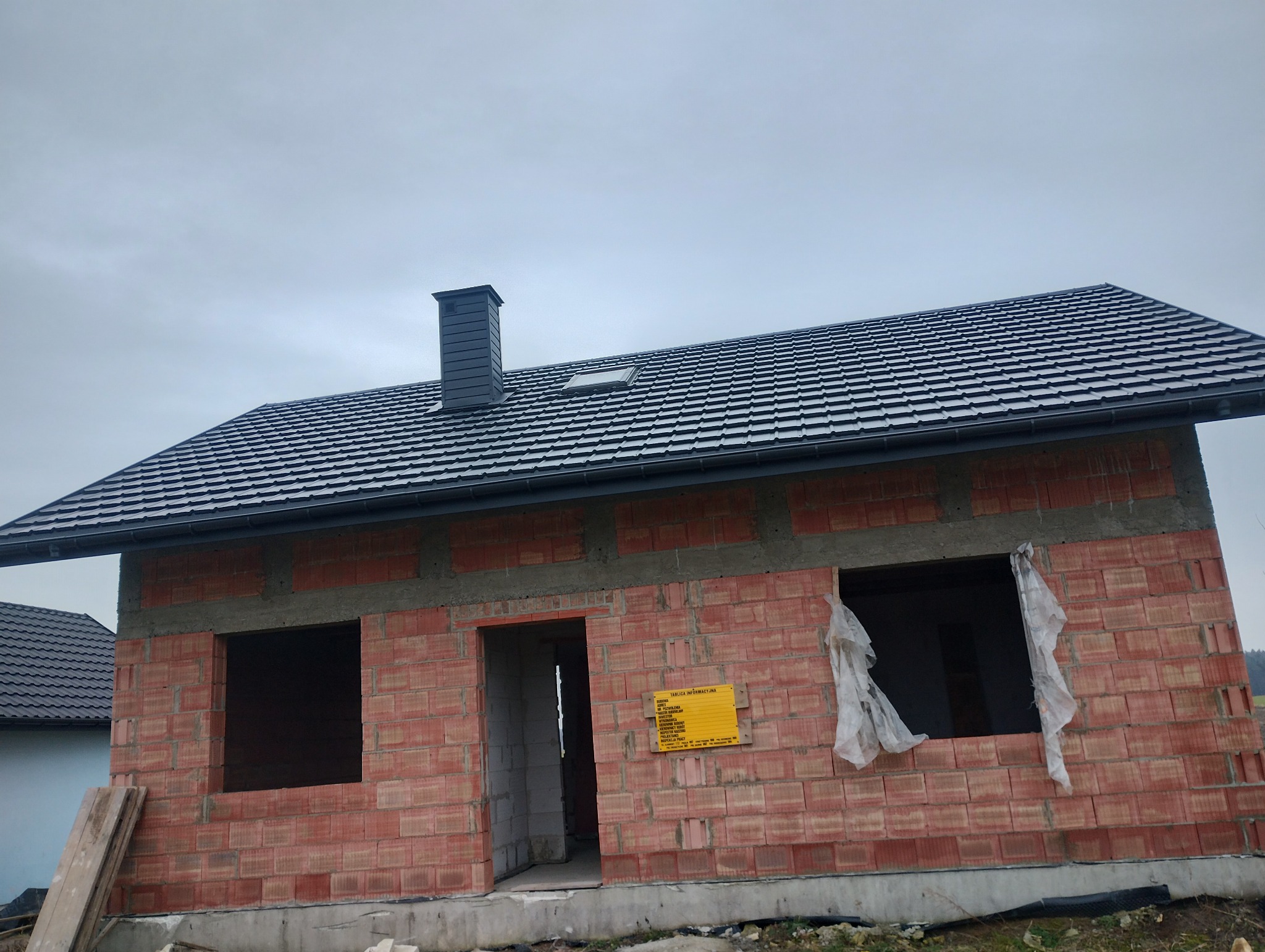 Surowy stan domu z czerwonej cegły z czarnym dachem i kominem, widoczna tablica informacyjna na ścianie, okna i drzwi bez stolarki.