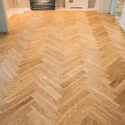 MP Parkiet Michał Piotrowski - Jasna, nowa podłoga drewniana ułożona w jodełkę, widok z góry. Fragment kolumny i drzwi w tle. Podłoga z wyraźnym rysunkiem słojów, ciepłe oświetlenie.