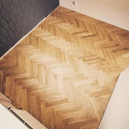 Ułożony parkiet w jodełkę klasyczną, z widocznym fragmentem grzejnika i ściany z wzorzystą tapetą w tle. Naturalne odcienie drewna i delikatne oświetlenie.