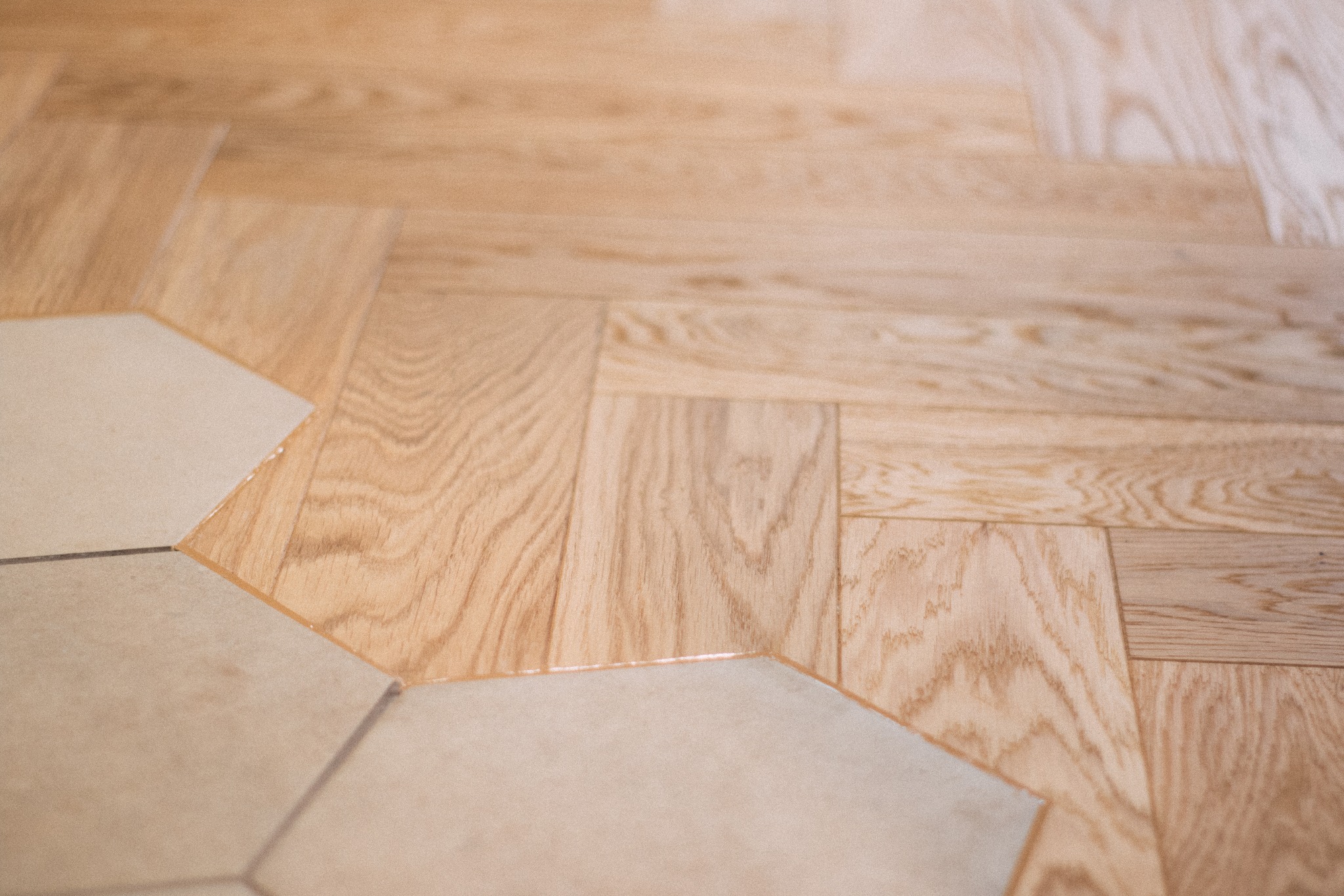 Jasny parkiet ułożony we wzór jodełki francuskiej, przechodzący w heksagonalne płytki podłogowe. Kontrast materiałów i faktur, ciepłe odcienie drewna i chłodne płytki.