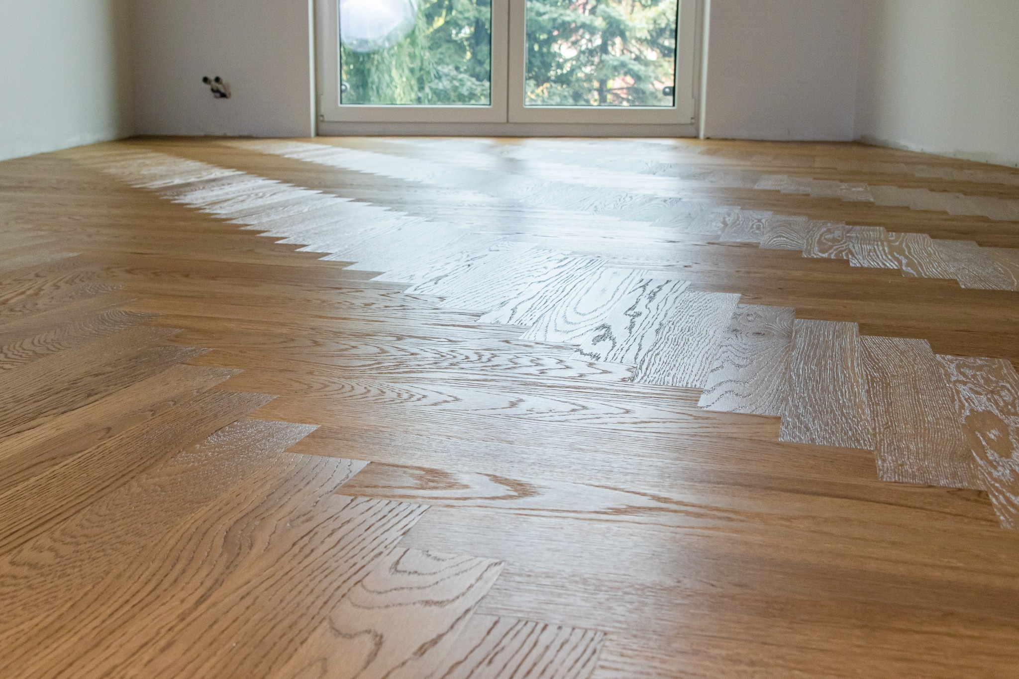 Ukończona podłoga z parkietu ułożonego w jodełkę, częściowo pokryta lakierem. Widoczne okno z zielenią w tle i fragment ściany z gniazdkiem.