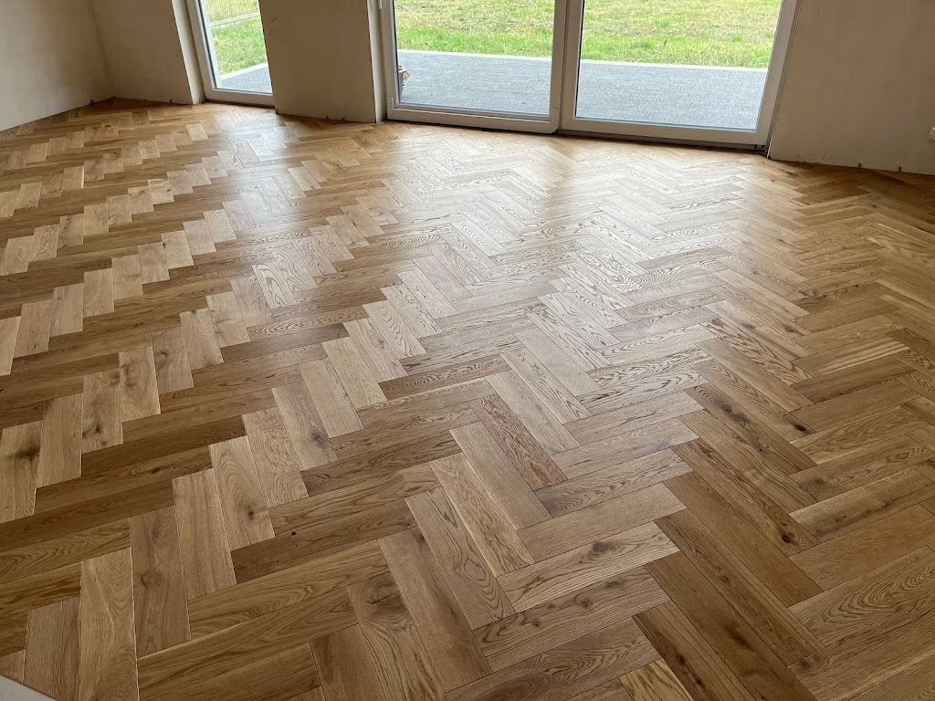 Jasna, dębowa podłoga ułożona w jodełkę francuską, widoczna w nowym, jasnym pomieszczeniu z dużymi oknami balkonowymi. Naturalne światło podkreśla strukturę drewna.