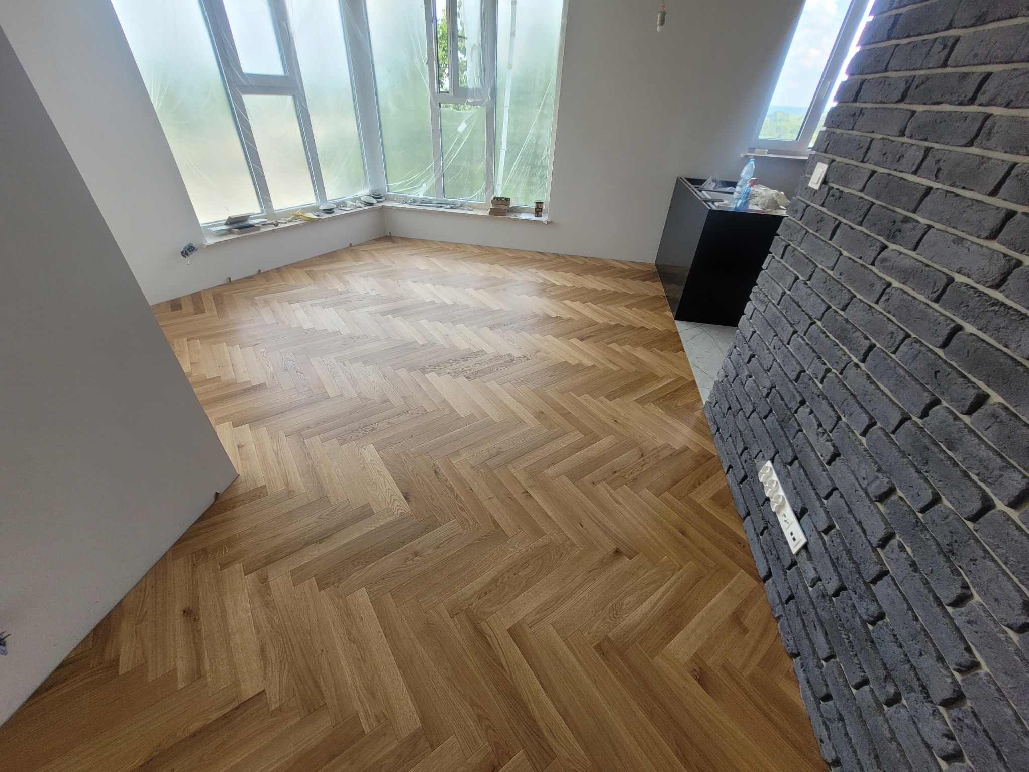 Ukończona podłoga z parkietu ułożonego w jodełkę w jasnym pomieszczeniu z oknem i fragmentem ściany z cegły. Widoczne gniazdka elektryczne.