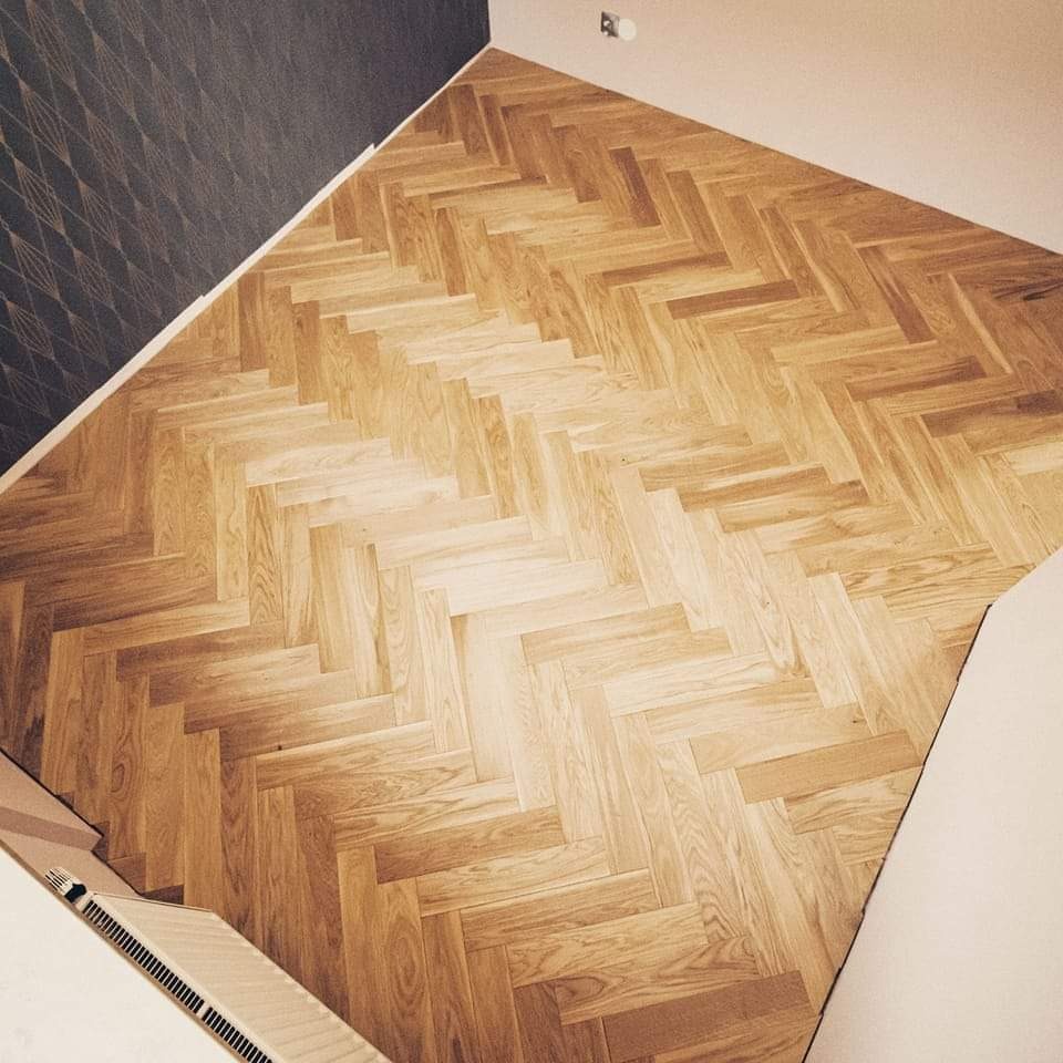 Ułożony parkiet w jodełkę klasyczną, z widocznym fragmentem grzejnika i ściany z wzorzystą tapetą w tle. Naturalne odcienie drewna i delikatne oświetlenie.