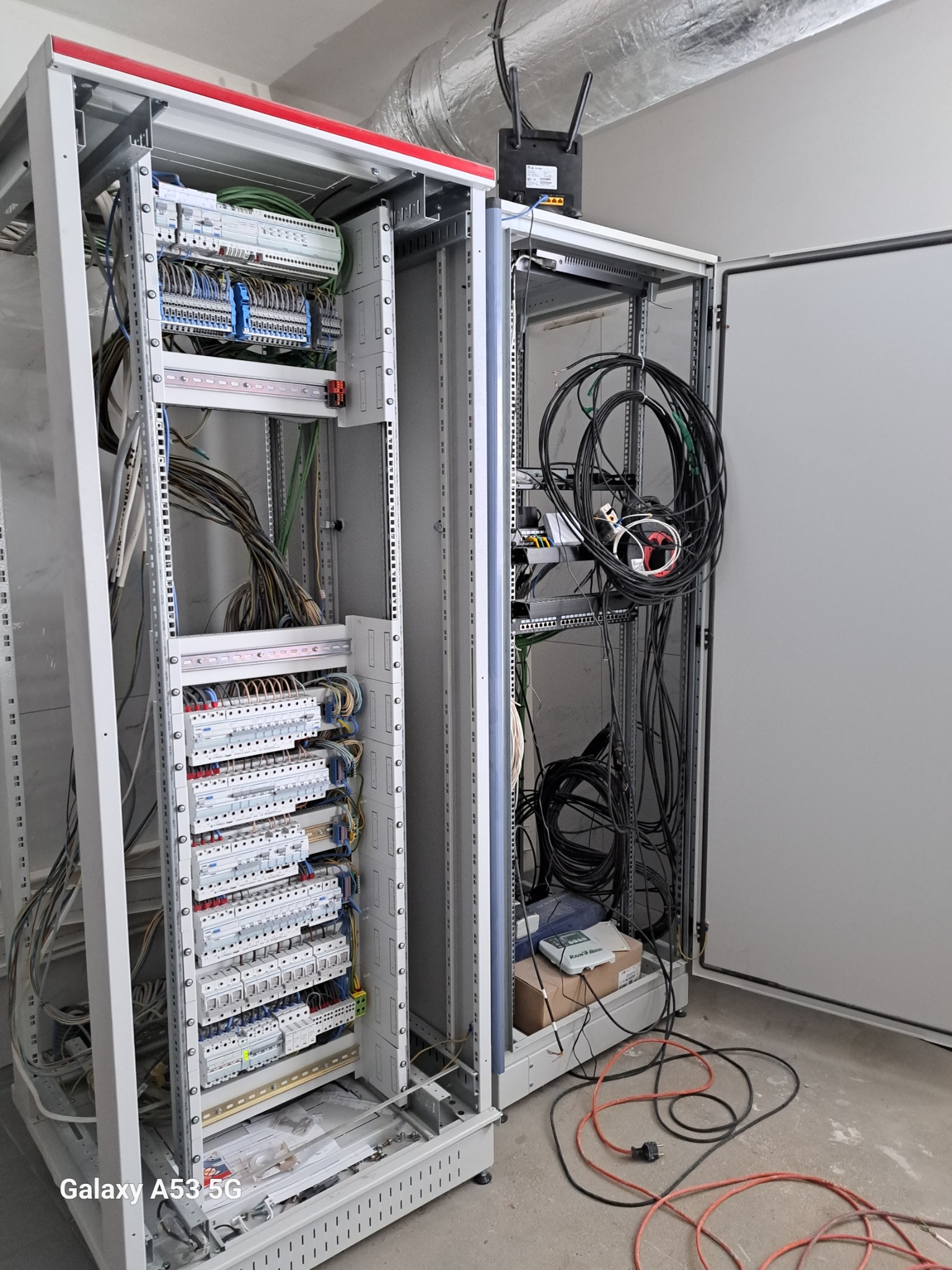 Dwie szafy elektryczne w trakcie montażu, widoczne liczne przewody, bezpieczniki i inne komponenty, na górze szafy router z antenami, na podłodze leżą kable zasilające.