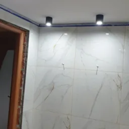 Wykończona ściana łazienki z białych płytek imitujących marmur, oświetlona trzema czarnymi lampami sufitowymi, widoczny fragment obudowy drzwi.