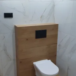 Nowoczesna toaleta podwieszana z drewnianą obudową stelaża, biała ceramika, marmurowe płytki ścienne i podłoga imitująca drewno.