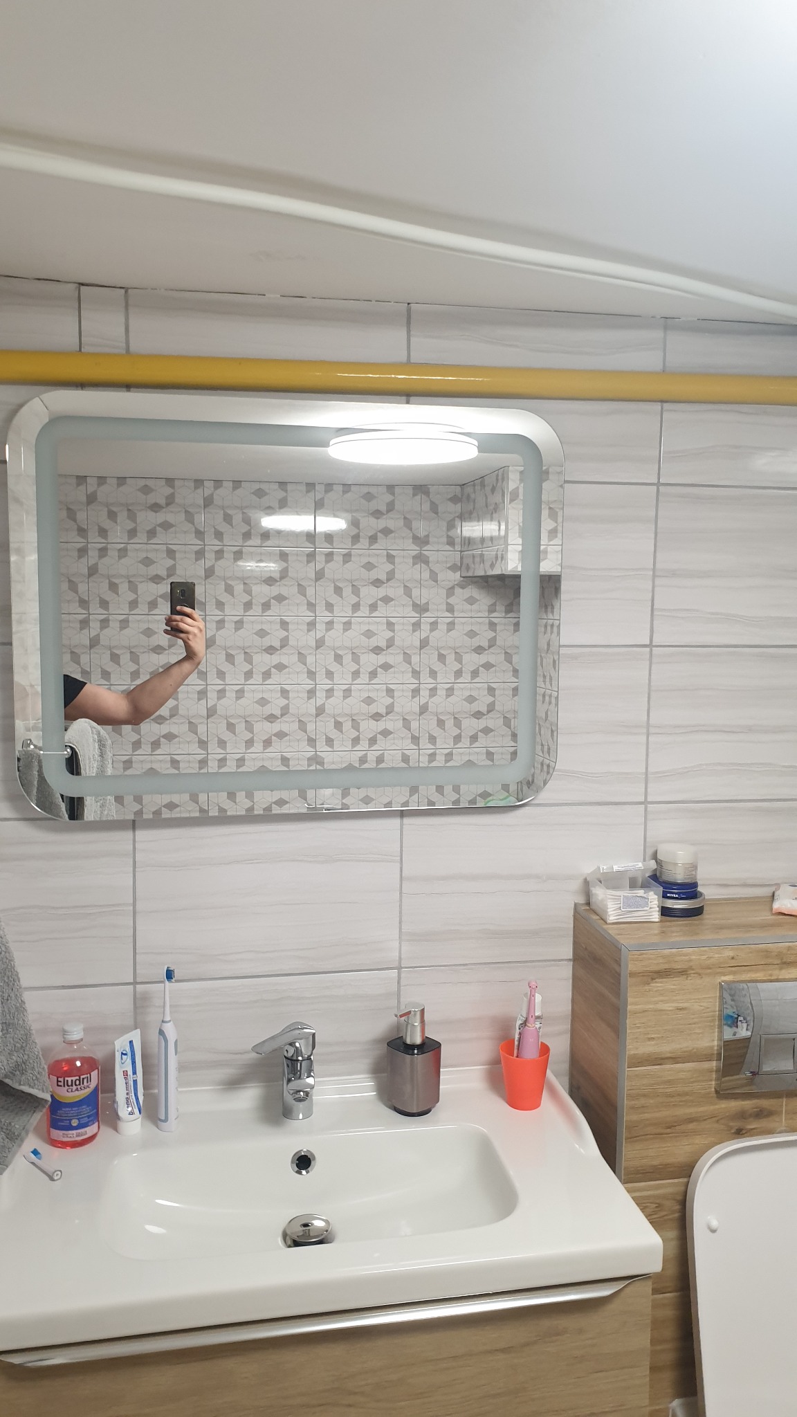 Umywalka z baterią, dozownikiem mydła i akcesoriami do higieny osobistej w łazience z mozaikową ścianą odbijającą się w podświetlanym lustrze, widoczny fragment instalacji gazowej pod sufitem.