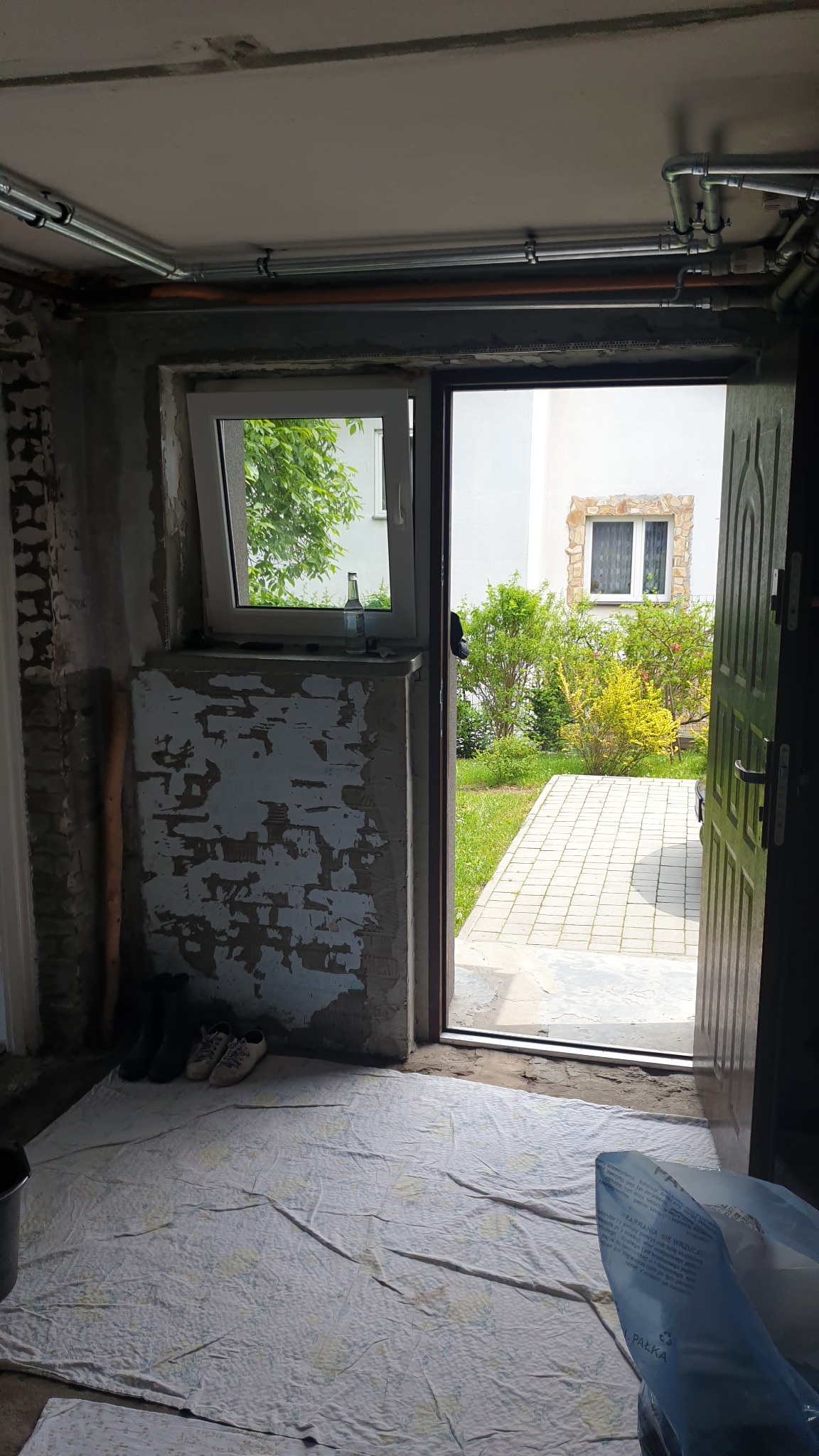 Wnętrze w trakcie remontu: widoczne niedokończone ściany z odpadającym tynkiem, zabezpieczona podłoga, otwarte drzwi na zewnątrz z widokiem na ogród i budynek.