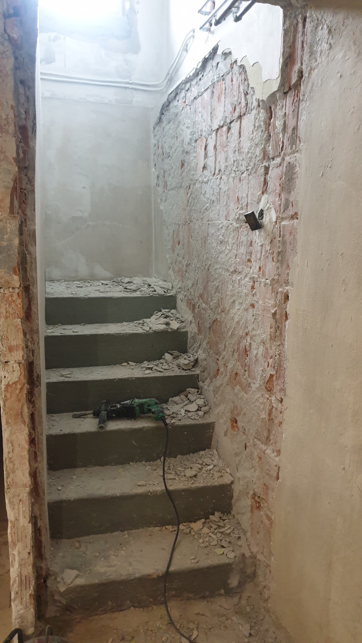 Schody w trakcie remontu: surowe betonowe stopnie pokryte gruzem, ściana z odsłoniętą cegłą i resztkami tynku, zielony młot udarowy na jednym ze stopni.