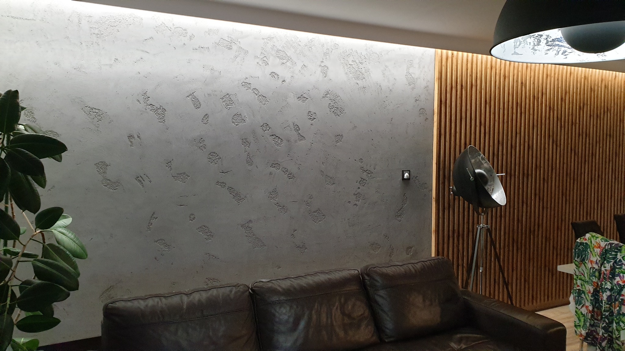 Szara ściana z dekoracyjną fakturą, oświetlona listwą LED, z fragmentem drewnianej boazerii, lampą studyjną i skórzaną sofą