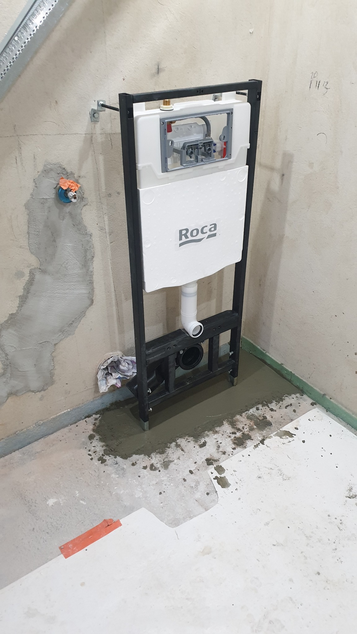 Stelaż podtynkowy WC marki Roca zamontowany w surowej łazience, widoczne przyłącza wodne i kanalizacyjne, fragment ściany z niedokończonymi pracami tynkarskimi.