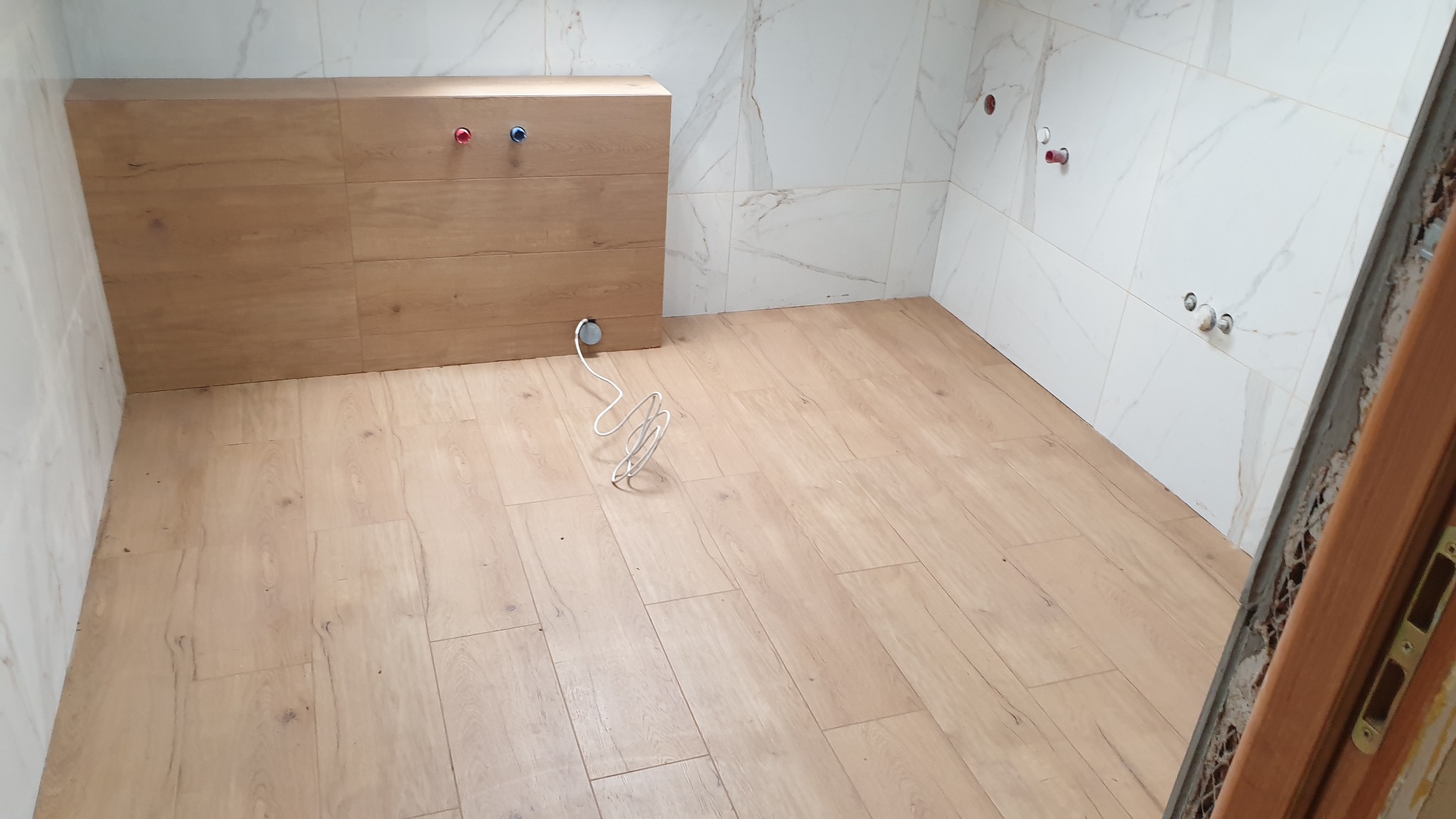 Nowa łazienka z drewnopodobną podłogą i marmurowymi płytkami ściennymi, widoczne wyprowadzenia instalacji wodnej i meble pod umywalkę z drewnopodobnym frontem