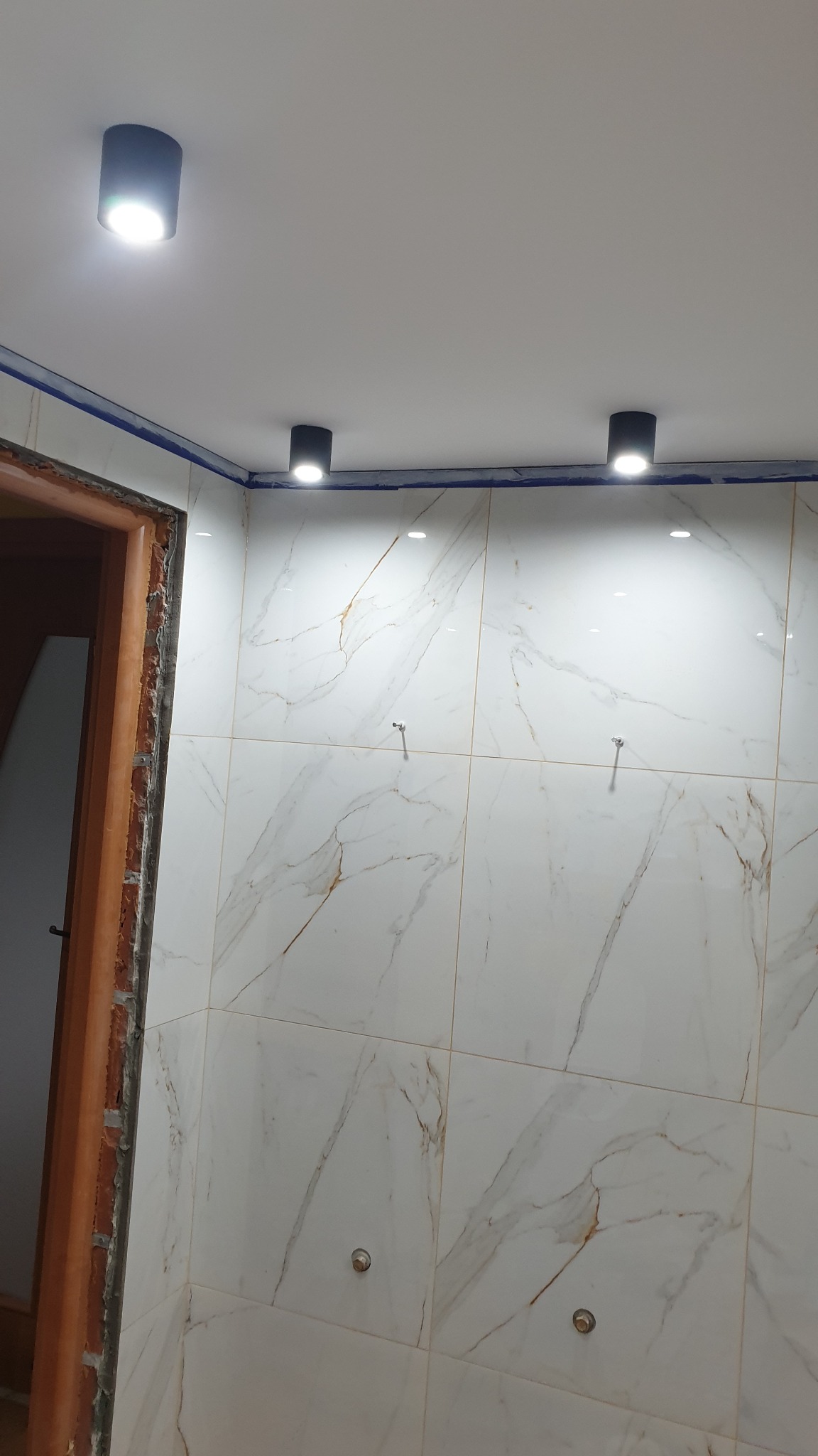 Wykończona ściana łazienki z białych płytek imitujących marmur, oświetlona trzema czarnymi lampami sufitowymi, widoczny fragment obudowy drzwi.