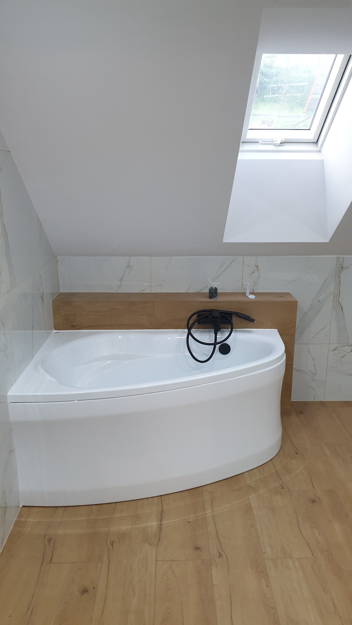 Nowoczesna biała wanna narożna z czarną armaturą zamontowaną na ścianie, drewniana półka nad wanną, marmurowe płytki ścienne, podłoga z paneli imitujących drewno, okno dachowe w skosie.