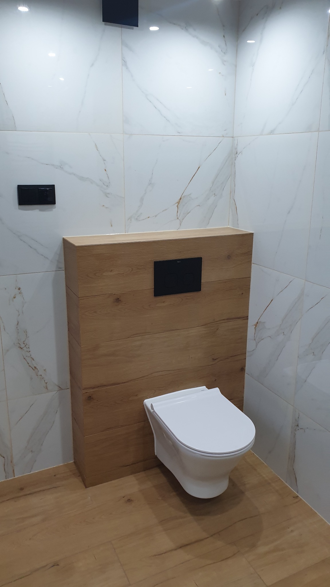 Nowoczesna toaleta podwieszana z drewnianą obudową stelaża, biała ceramika, marmurowe płytki ścienne i podłoga imitująca drewno.