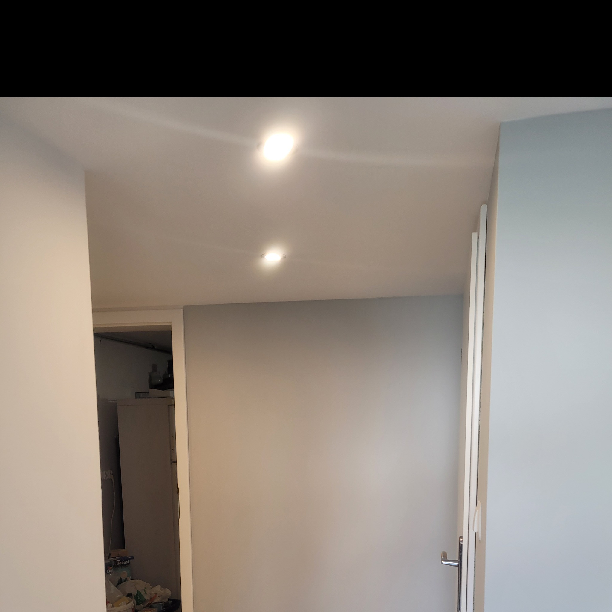 Świeżo pomalowane wnętrze z widocznymi dwoma punktowymi lampami sufitowymi i otwartymi drzwiami do zagraconego pomieszczenia.