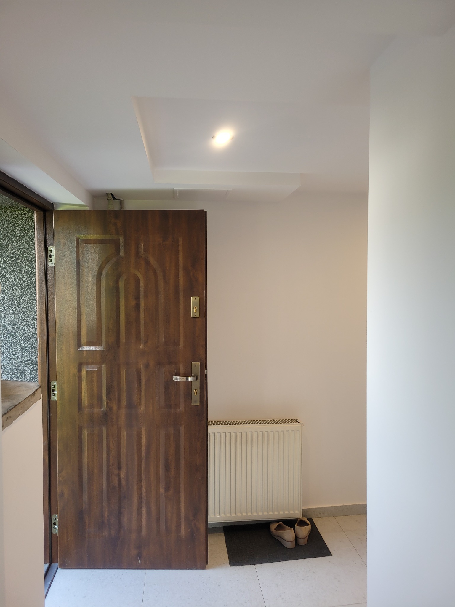 Brązowe, drewniane drzwi wejściowe z dekoracyjnymi frezowaniami, otwarte do wnętrza, widoczny biały grzejnik i buty na wycieraczce, minimalistyczne wnętrze.