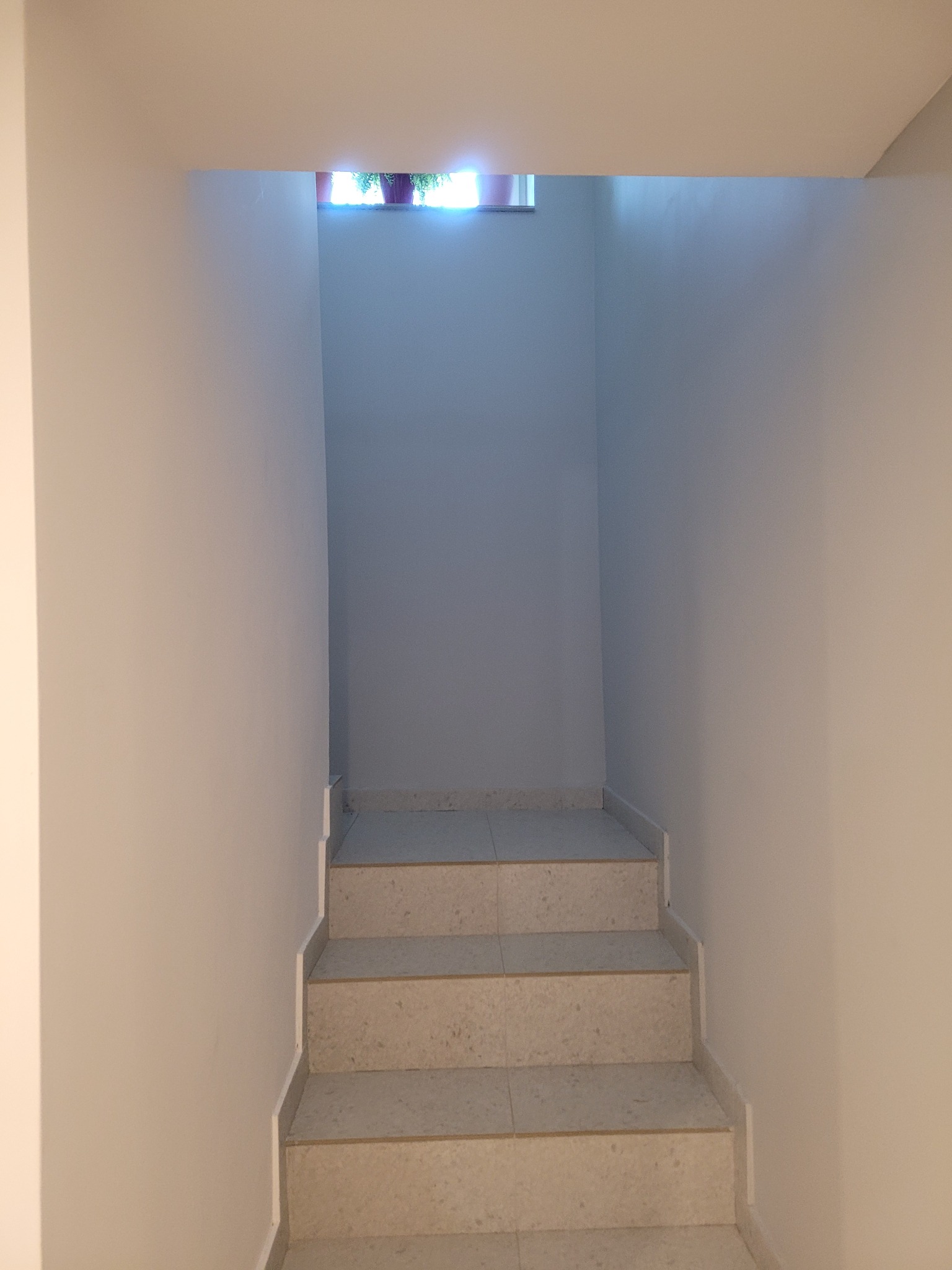 Klatka schodowa z jasnymi, marmurkowymi schodami prowadząca w górę, widoczne doniczki z roślinami w oknie na końcu schodów.