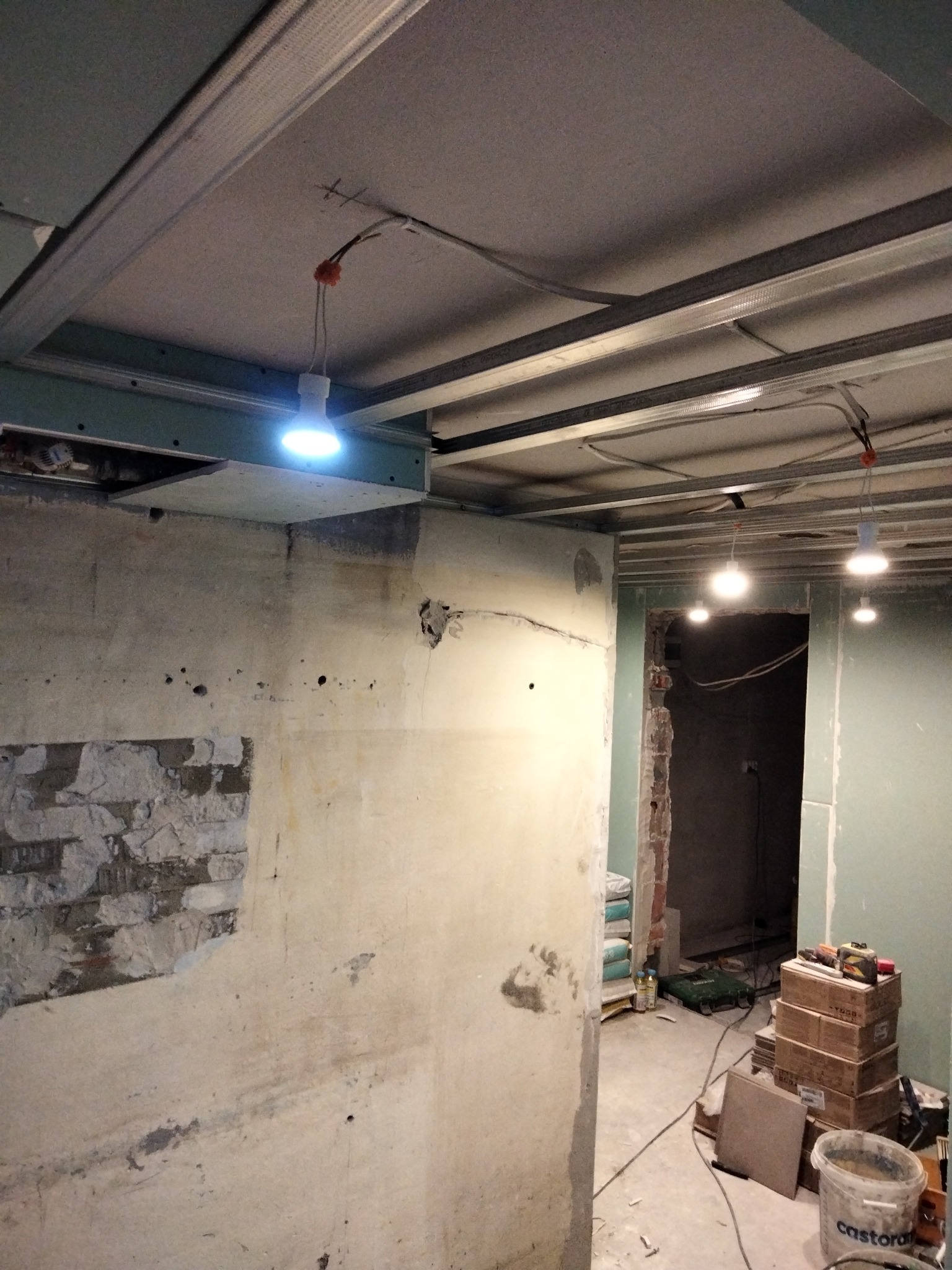 Wnętrze w trakcie prac wykończeniowych: surowa ściana z fragmentami cegieł, metalowa konstrukcja sufitu podwieszanego z widocznym oświetleniem roboczym, w tle materiały budowlane.