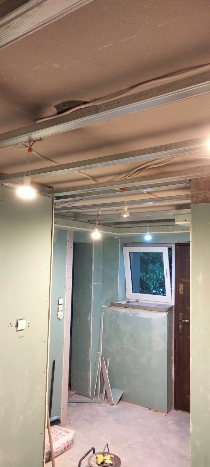 Wnętrze w trakcie montażu płyt gipsowo-kartonowych na ścianach i suficie, widoczne metalowe profile konstrukcyjne, tymczasowe oświetlenie i worki z zaprawą na podłodze.