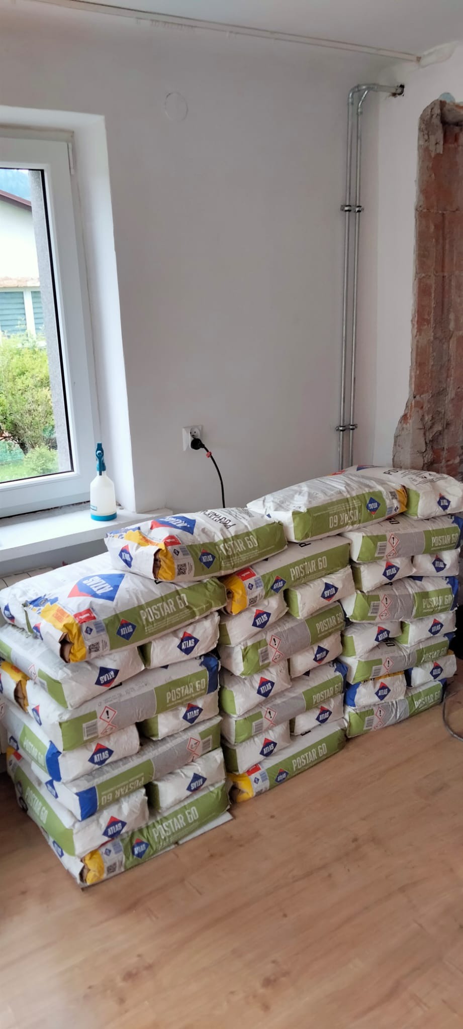 Stos worków zaprawy Atlas Postar 60 w pomieszczeniu w trakcie remontu, widoczne instalacje elektryczne i fragment ściany z odsłoniętą cegłą