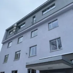 Szary budynek wielorodzinny z widocznymi oknami, balkonem i trzema klimatyzatorami na elewacji, sfotografowany pod kątem podczas lekkiego opadu śniegu.