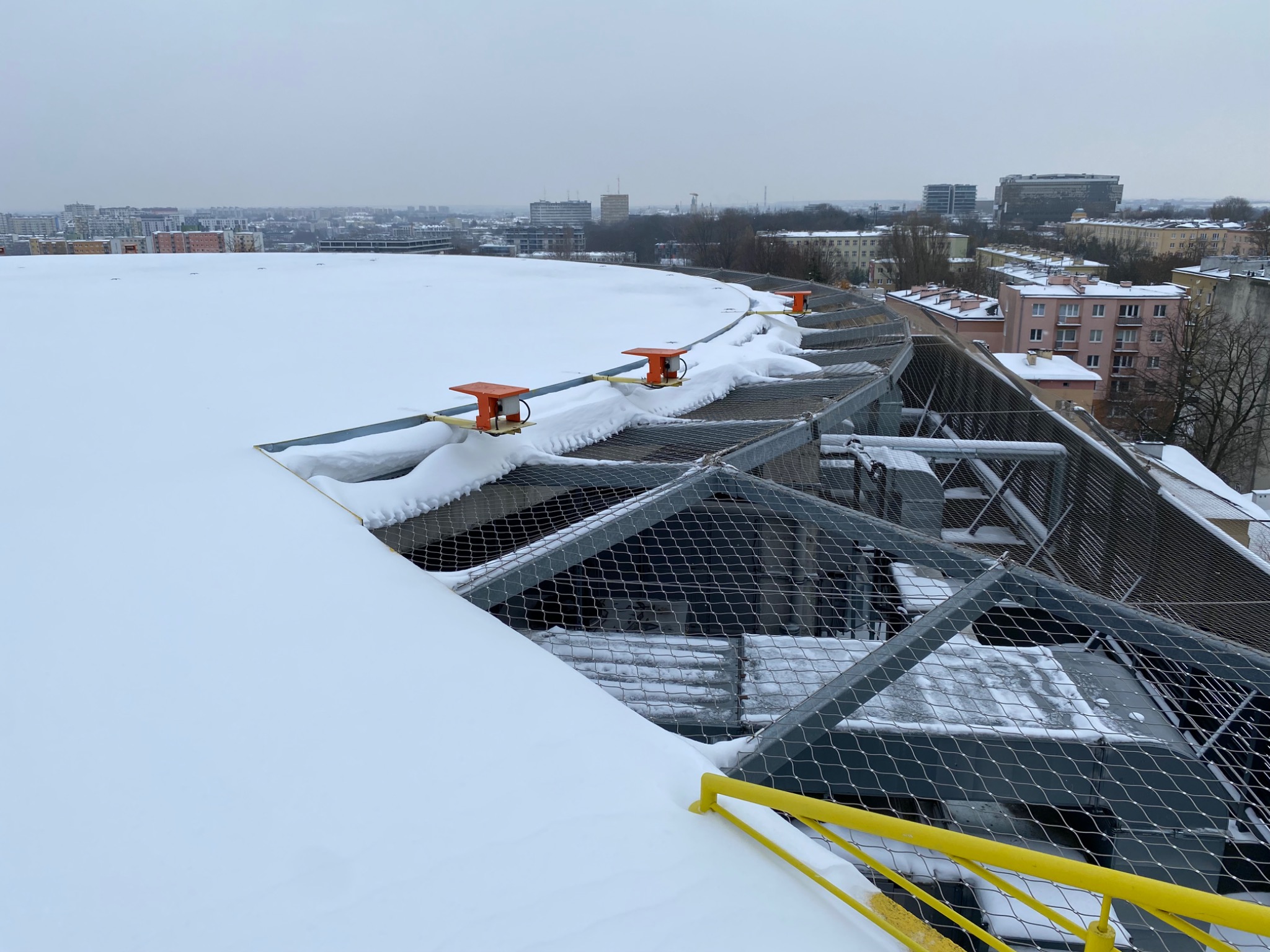 Zabezpieczenie dachu siatką przeciwśniegową z pomarańczowymi uchwytami, widok na zasypane śniegiem miasto w tle, pochmurny dzień.