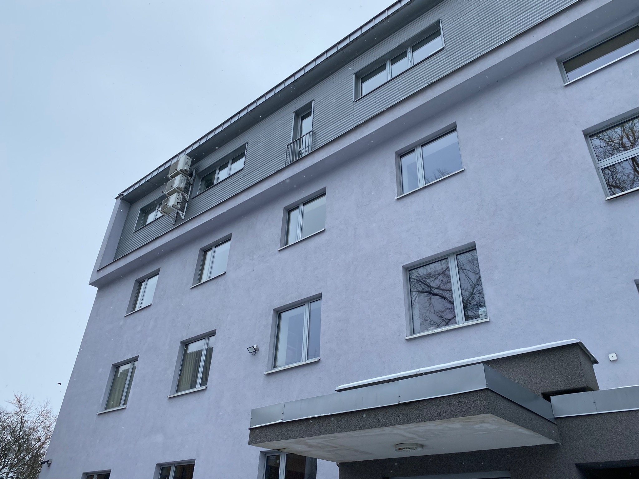 Szary budynek wielorodzinny z widocznymi oknami, balkonem i trzema klimatyzatorami na elewacji, sfotografowany pod kątem podczas lekkiego opadu śniegu.
