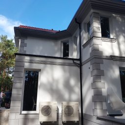 L.i.n.budservis - Elewacja domu z dekoracyjnymi elementami, dwoma klimatyzatorami Mitsubishi, oraz rzeźbą głowy na tle błękitnego nieba i drzew