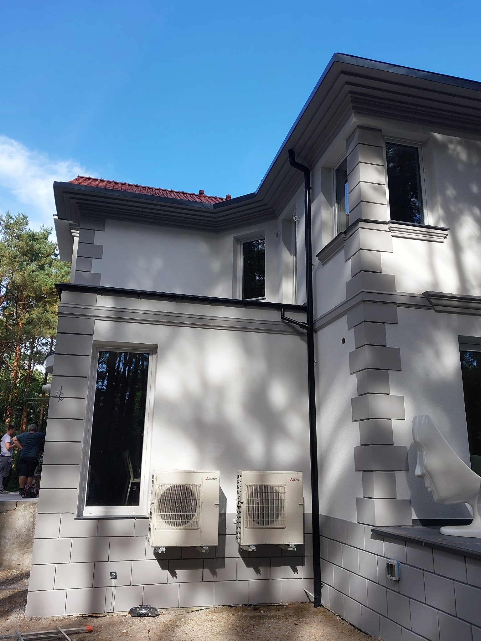 Elewacja domu z dekoracyjnymi elementami, dwoma klimatyzatorami Mitsubishi, oraz rzeźbą głowy na tle błękitnego nieba i drzew