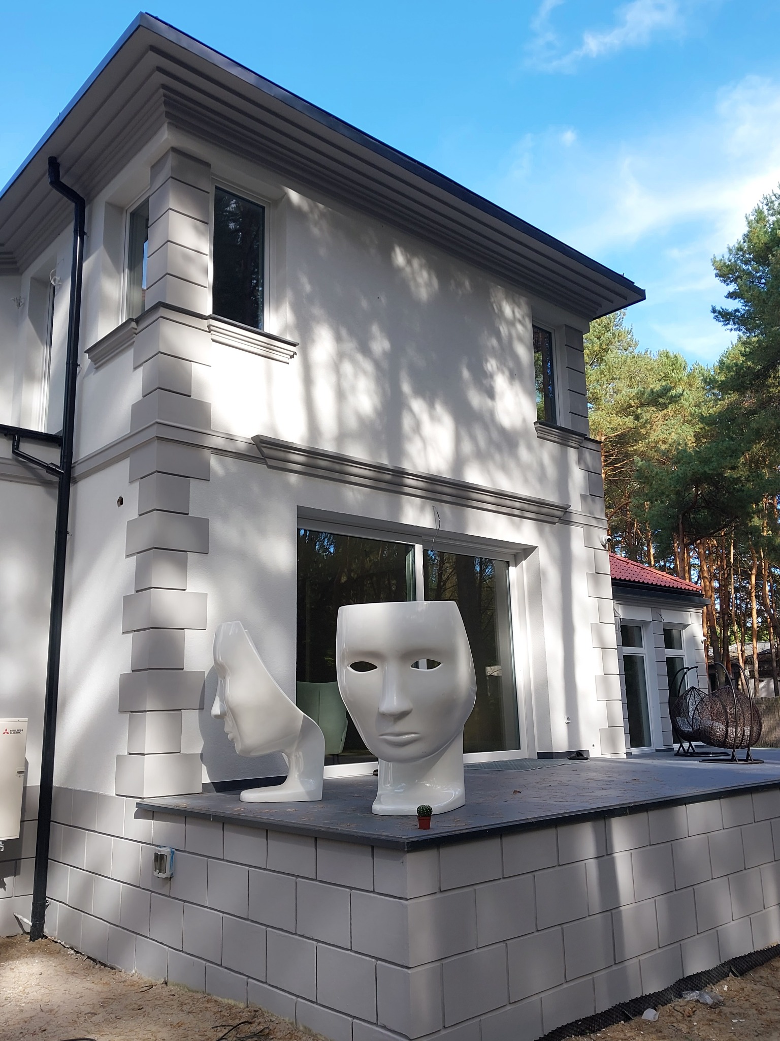 Elewacja domu jednorodzinnego z dekoracyjnymi elementami w odcieniach szarości, dwie białe rzeźby w kształcie głowy na tarasie, widok na las w tle.