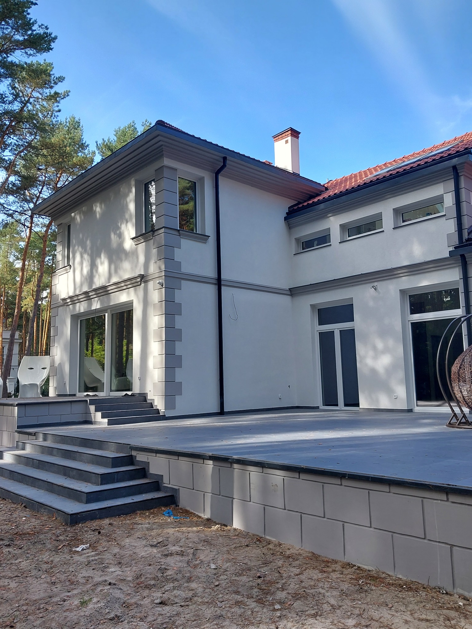 Nowoczesny dom jednorodzinny z białymi ścianami i szarymi elementami dekoracyjnymi elewacji, widok z zewnątrz na taras i schody wejściowe.