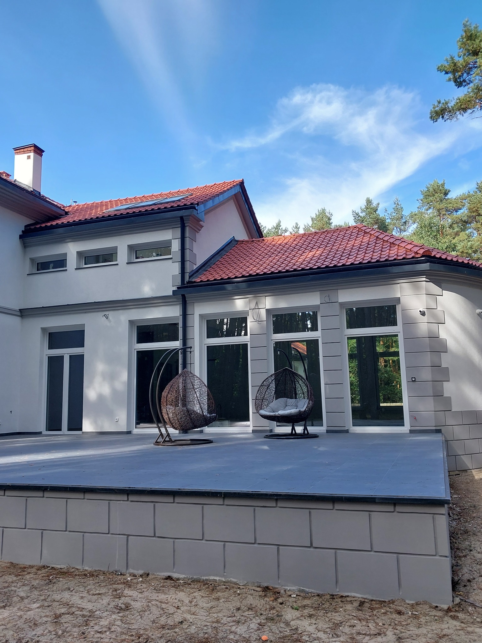 Nowoczesny dom z szarą elewacją, czerwonym dachem i tarasem z dwoma wiszącymi fotelami typu kokon