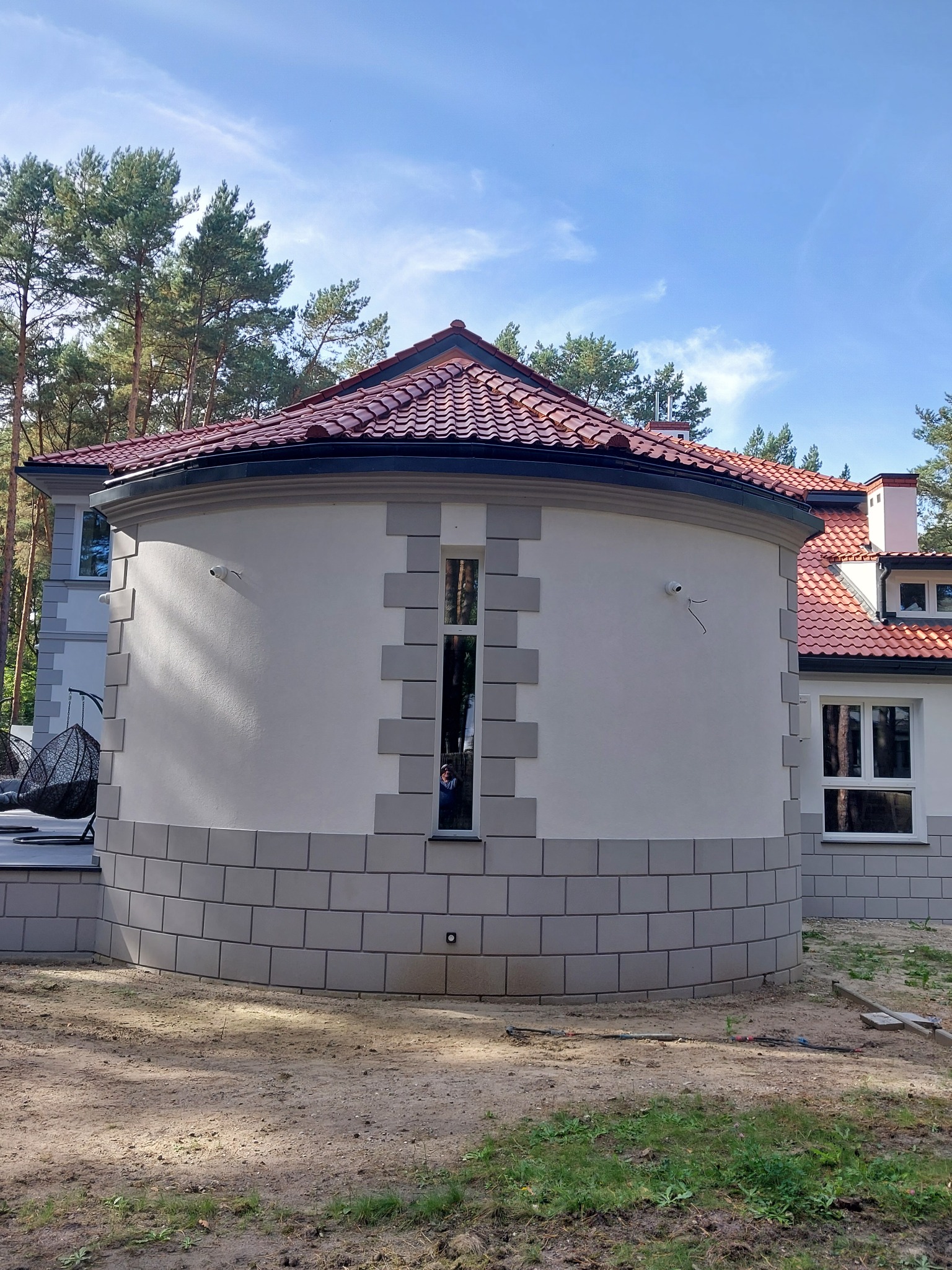 Zaokrąglona część domu z jasną elewacją i szarym cokołem z imitacji kamienia, widoczna z zewnątrz na tle drzew i nieba.